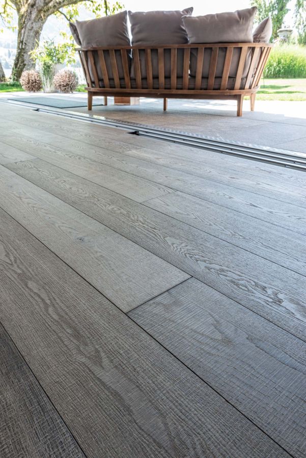 Detailaufnahme: Landhausdiele grau-braun, sägerau. Im Hintergrund ist die Gartenlounge mit voluminösen Kissen sichtbar.